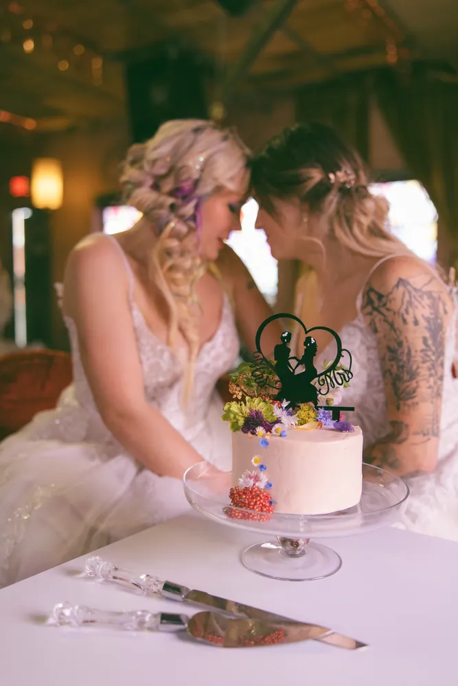 Two brides kiss near a wedding cake with floral accents, celebrating their marriage in a warmly lit venue.