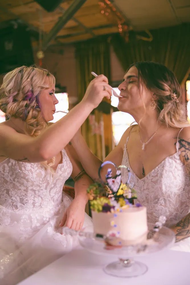 Two brides share a tiered cake, smiling, at a wedding celebration. Floral details adorn the cake and hair.