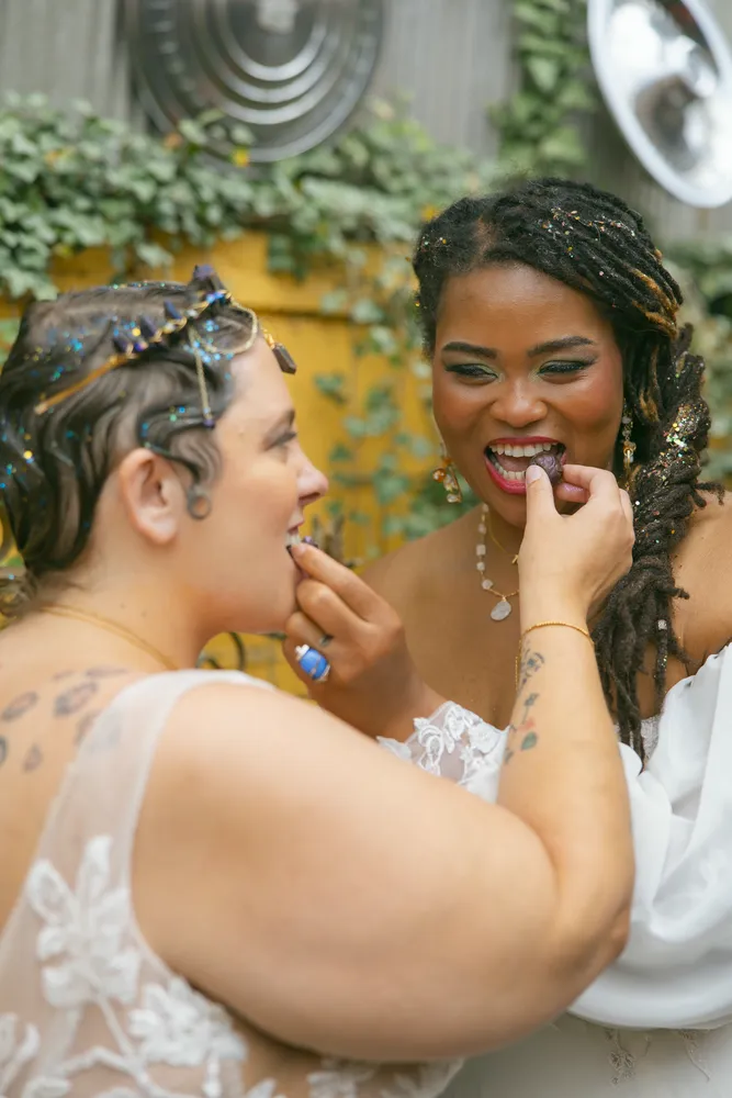 A person with dark hair and a white dress receives a hug from another person, both smiling with glitter on their faces.