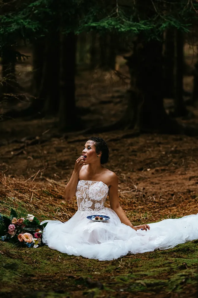 Woman in a wedding dress sits in a forest, holding a small cake, surrounded by flowers.