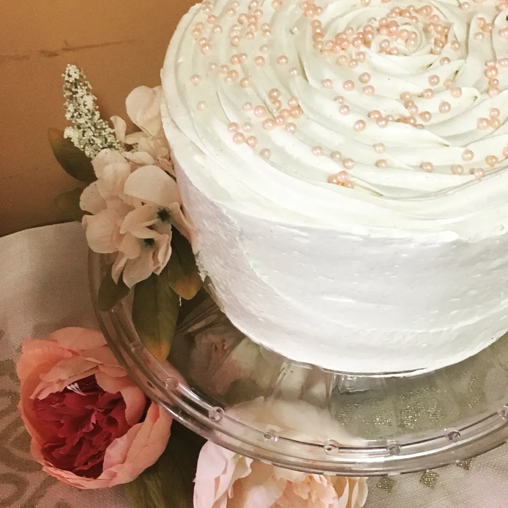 Elegant white cake on a glass stand, decorated with fresh flowers and edible pearl accents. Perfect for a special occasion.