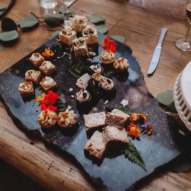 Assorted miniature desserts on a slate platter, featuring mousse tarts and flower garnishes. A knife rests nearby.