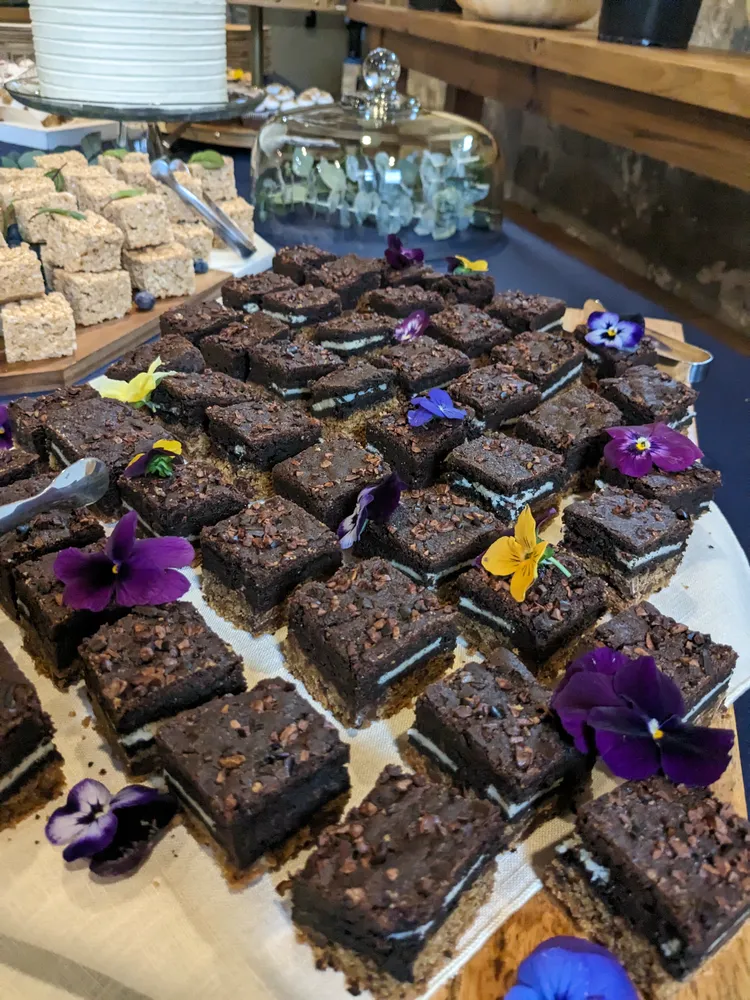 Chocolate brownies topped with edible flowers sit on a wooden board, alongside a tiered display of coconut macaroons.