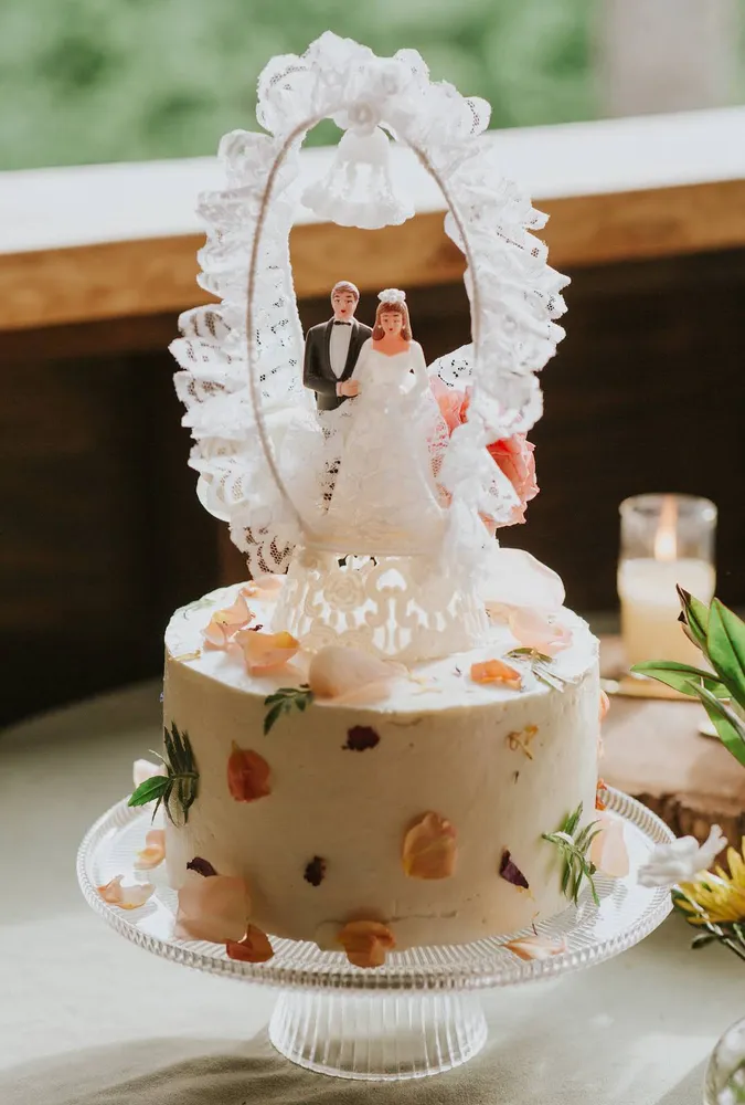 Tiered wedding cake with a bride and groom cake topper, decorated with fruit and flowers on a decorative stand.