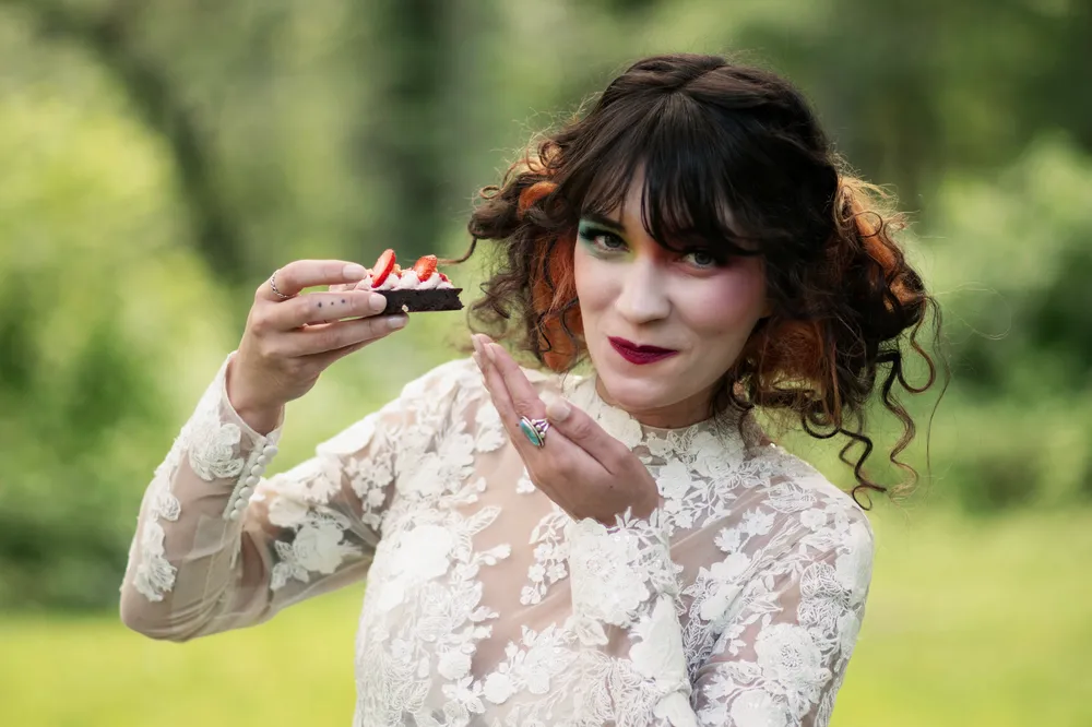 Person holding a strawberry cake slice, smiling, with curly red hair and elaborate eye makeup, wearing a lace dress.