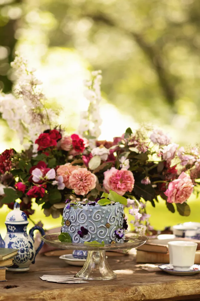 Layered cake on a pedestal, adorned with flowers and blue icing, sits on a table with vintage china.