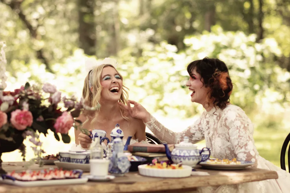Two women share a table laden with food and flowers during an outdoor picnic in a garden.